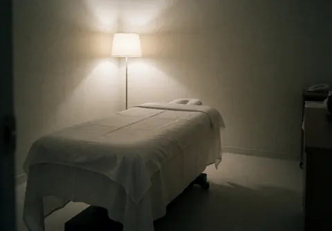 Lit de massage recouvert de draps blancs dans une pièce aux murs neutres, éclairé par une lampe sur pied, idéal pour les massages cervicaux.