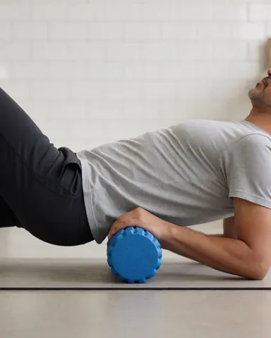 Un homme pratique un massage des tissus profonds avec un rouleau bleu sur un tapis de yoga dans un gymnase aux murs carrelés blancs.