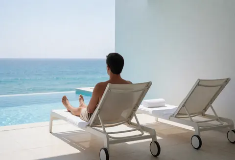 Un homme se repose sur un transat blanc à côté d'une piscine à débordement qui semble se fondre avec l'océan, avec des murs blancs et un autre transat vide à ses côtés.