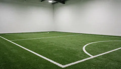 Vue d'un terrain de football en intérieur avec des lignes blanches distinctes, des murs blancs et un éclairage clair, idéal pour un club sportif.