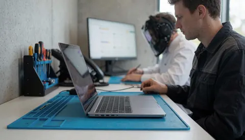 Un technicien répare un ordinateur portable sur une station de travail équipée d'outils et d'un moniteur, dans un espace avec murs en béton offrant un espace pour le texte.