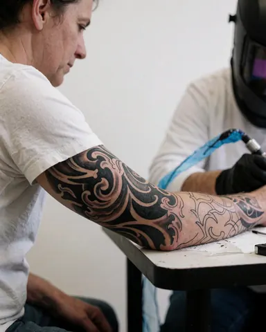 Tatoueur travaillant sur un bras avec un tatouage en cours, mur blanc, machine visible, client assis.