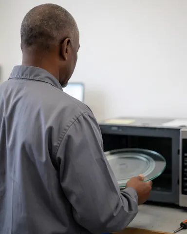 Technicien de réparation installant un nouveau plateau en verre dans un micro-ondes ouvert au sein d'un atelier aux murs blancs.
