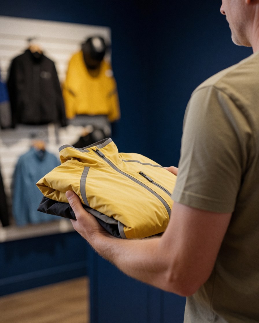 Cliente tenant une veste jaune avec détails réfléchissants dans un magasin de vélo, fond avec vêtements cyclistes sur mur bleu foncé.