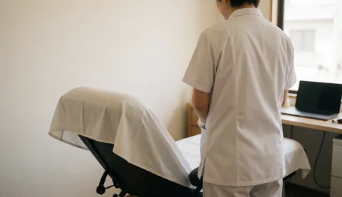 Professionnel en uniforme blanc de dos, à côté d'un fauteuil inclinable recouvert d'un tissu blanc dans une salle de thérapie lumineuse aux murs clairs.