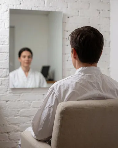 Une cliente assise observe son reflet sain dans un miroir, avec un professionnel en uniforme blanc en arrière-plan, dans un cadre calme.