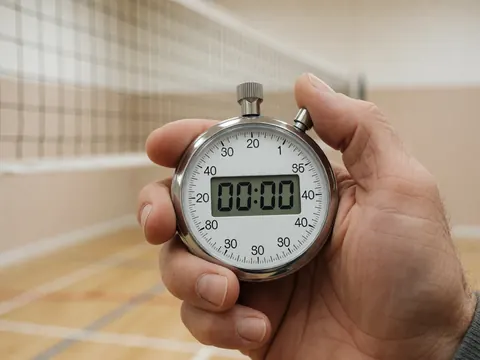 Main tenant un chronomètre numérique affiché à zéro devant un filet de volleyball dans un gymnase, idéal pour un entraîneur de sport collectif.