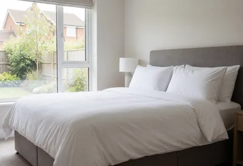 Lit king-size avec draps blancs et oreillers, face à une fenêtre offrant une vue sur le jardin, dans une chambre moderne et propre d'une maison de vacances.