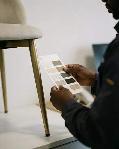 Une main tenant une carte de couleurs devant une chaise dorée dans un magasin de meubles, sur un fond blanc.