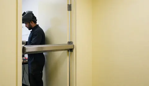 Un technicien de sécurité installe une barre magnétique sur une porte de bureau avec des murs jaunes et des outils visibles, dans le cadre d'une réparation d'alarme professionnelle.