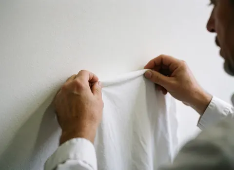 Mains d'un opérateur vérifiant la qualité du tissu d'un t-shirt blanc contre un mur blanc, détail de la production textile.