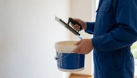 Un travailleur en uniforme bleu tient un seau d'adhésif et une truelle, prêt à installer un sol en liège sur un mur blanc éclairé par la lumière naturelle.