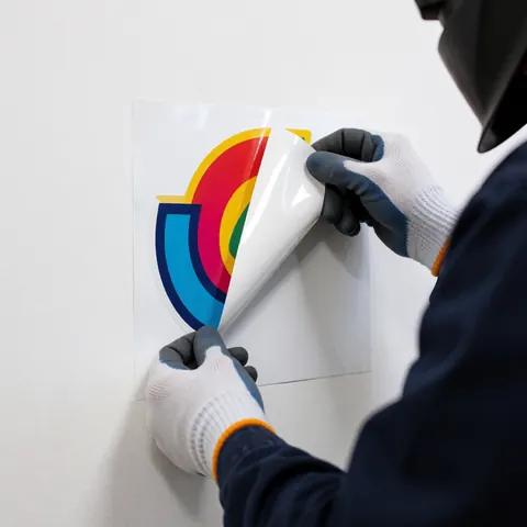 Des mains gantées appliquent un vinyle coloré à motif graphique sur un mur blanc propre, illustrant un service d'impression professionnelle.