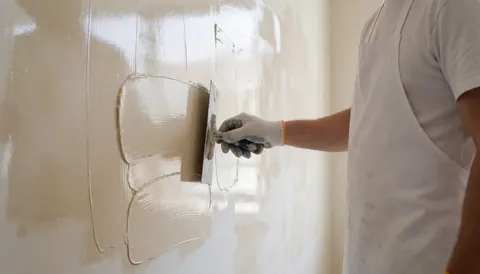 Un peintre professionnel applique un enduit vénitian brillant sur un mur blanc à l'aide d'une truelle, portant des gants et un tablier blancs.