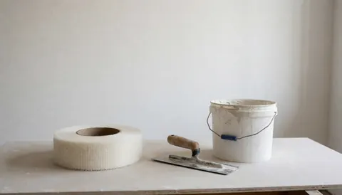 Rouleau de treillis de verre, pelle et seau de mastic posés sur un panneau de plâtre, outils pour finitions de murs blancs.