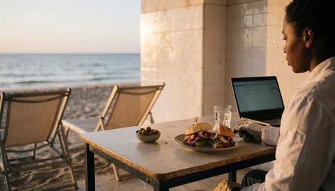 Personne travaillant sur ordinateur portable à une table de plage avec sandwich de poulpe, olives et vue sur la mer.