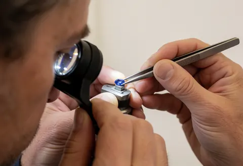 Main utilisant des pinces pour inspecter un saphir bleu avec une loupe sur un support métallique, fond blanc, pour la réparation de bijoux.