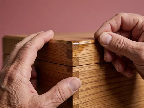 Gros plan de mains appliquant du vernis sur une jonction de bois avec biseau, détail de réparation de meubles sur fond rose