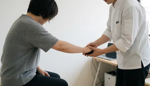 Un thérapeute guide la main d'un client tenant une balle de massage dans une clinique aux murs blancs et à la lumière naturelle.
