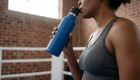 Une femme boit de l'eau d'une bouteille bleue sur un ring de boxe avec un mur en briques et une lumière naturelle, idéal pour un site web d'instructrice de boxe.