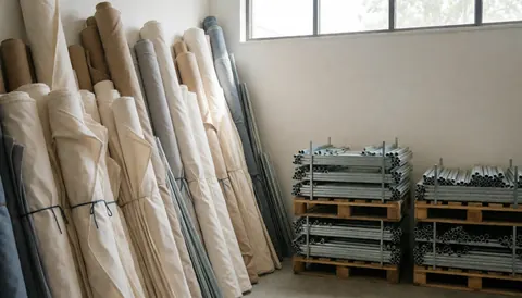 Entrepôt de stores avec rouleaux de tissu et paquets de tubes métalliques sous lumière naturelle, murs blancs et sol propre.