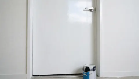 Porte blanche fraîchement peinte avec un seau de peinture synthétique sur le seuil, murs blancs et reflet du ciel, idéal pour les peintres en bâtiment.