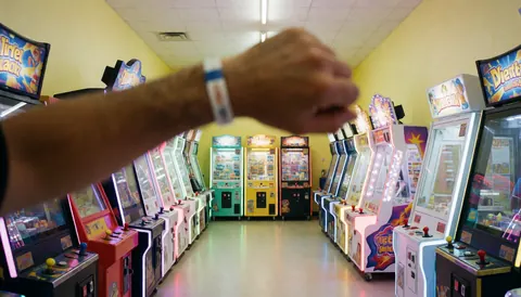 Premier plan d'une main avec un bracelet, couloir de machines arcade aux lumières vives et murs jaunes, idéal pour une page d'accueil de centre de loisirs.