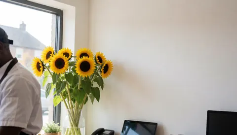 Un vase rempli de tournesols posé près d'une fenêtre, mur blanc et espace pour texte, silhouette travaillant dans une fleuristerie.