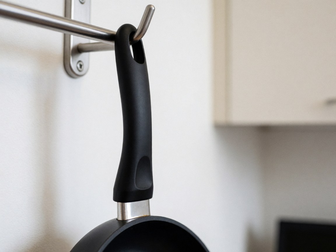Poêle de cuisine noire suspendue à un support métallique dans une cuisine aux murs blancs et aux armoires floues en arrière-plan.