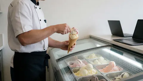 Vendeur de glace en uniforme blanc et noir servant un cône de glace fraîche depuis un chariot, sur fond propre et moderne.