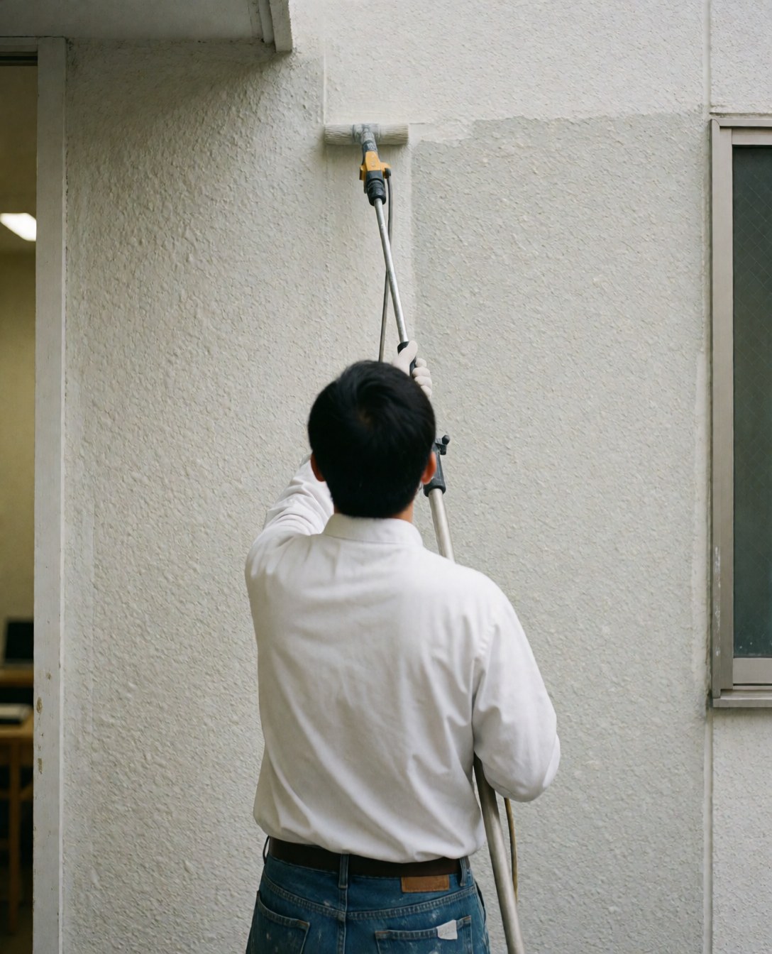 Un peintre applique un produit de protection sur un mur extérieur texturé à l'aide d'un rouleau avec une rallonge, vu de dos.