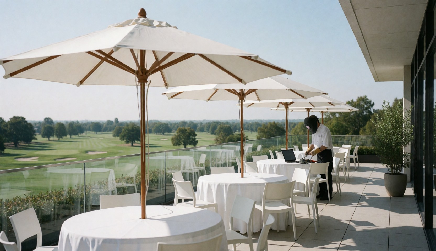 Terrasse avec tables rondes et parasols blancs offrant une vue panoramique sur le golf, une personne travaille sur place.
