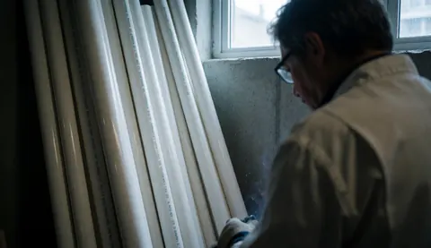 Un homme avec des lunettes manipule des tuyaux blancs en PVC dans un atelier au mur de béton éclairé par la lumière naturelle.