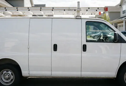Fourgon blanc équipé d'une échelle sur une rue résidentielle, prêt pour des interventions de chauffage, avec des reflets sur les vitres.