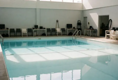 Piscine publique vide reflétant la lumière naturelle, chaises et tables rangées sur le deck, murs gris et propres.