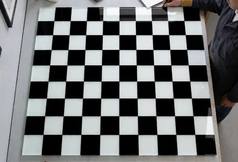 Panneau de verre décoratif avec motifs noirs et blancs, prêt à être installé dans un atelier de vitrerie aux murs gris avec des outils visibles.