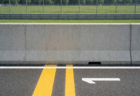 Barrage en béton avec lignes jaunes et flèche blanche sur piste de course, fond d'herbe et clôture métallique.