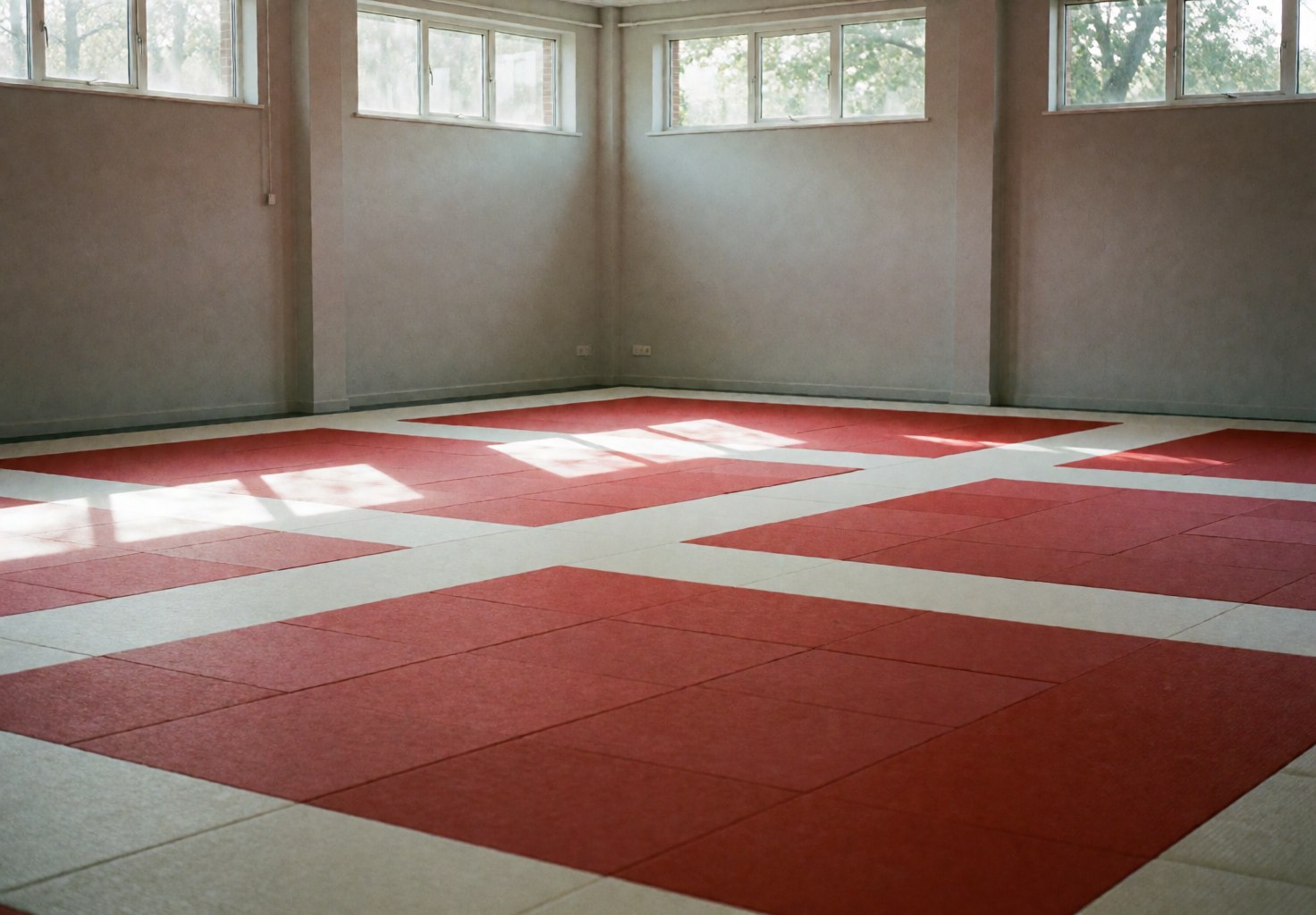Dojo de karate vide avec des tapis rouges et blancs, des murs gris et la lumière du soleil entrant par des fenêtres hautes, prêt pour l'entraînement.