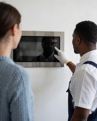 Un technicien en gants teste un micro-ondes encastré dans un mur blanc tandis qu'un client observe attentivement le nouvel appareil.