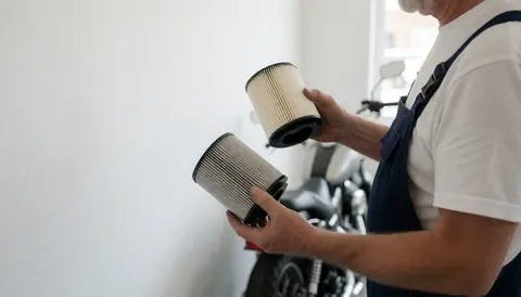 Mécanicien tenant un filtre neuf et un filtre sale devant une moto dans un atelier de réparation avec un mur blanc en arrière-plan.
