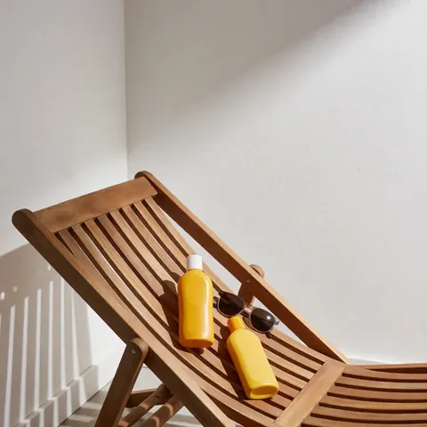 Chaises en bois disposées devant un mur blanc avec des bouteilles de crème solaire et des lunettes de soleil, créant une ambiance d'ombre et de détente idéale pour une maison de vacances.