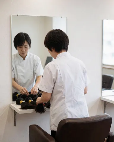 Un coiffeur range des extensions capillaires sur une étagère dans un salon moderne, devant un miroir et une chaise de coiffure.