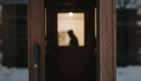 Porte en bois sombre éclairée par une lumière chaude à l'intérieur, avec de la neige à l'extérieur, idéale pour un fond de texte dans un refuge de montagne.