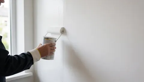 Un peintre applique de la peinture blanche sur un mur avec un rouleau, laissant un espace vide pour le texte, éclairé par la lumière naturelle d'une fenêtre.