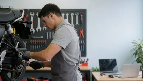 Mécanicien portant des lunettes et un tablier ajuste la chaîne d'une moto dans un atelier. Des outils sont suspendus au mur, dans un environnement professionnel et propre.