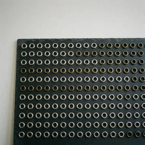 Rangées d'oeillets métalliques argent et bronze ancien disposés sur une surface d'ardoise sombre, adaptés à l'industrie textile ou du cuir