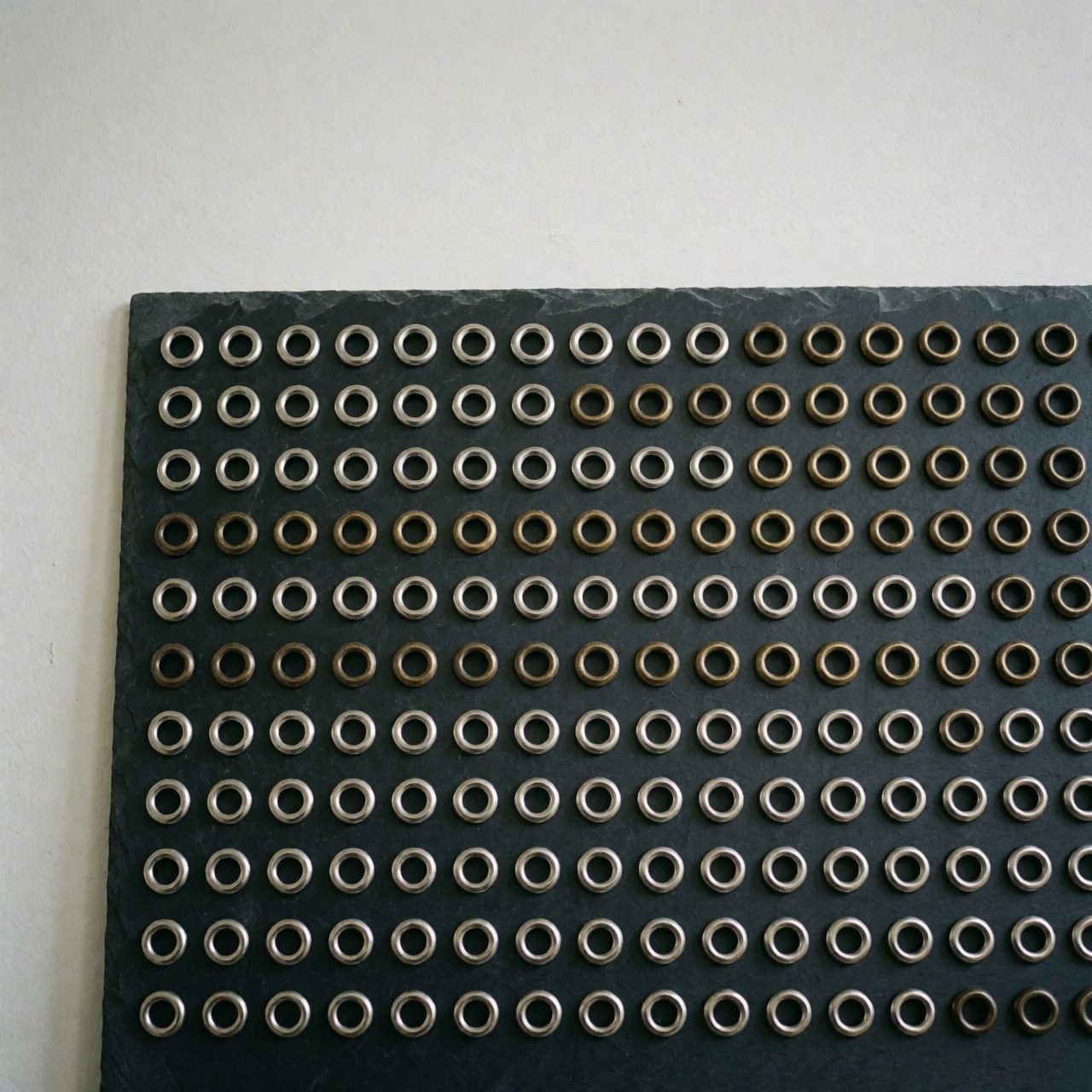 Rangées d'oeillets métalliques argent et bronze ancien disposés sur une surface d'ardoise sombre, adaptés à l'industrie textile ou du cuir
