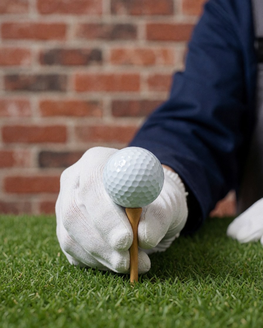 Une main portant un gant blanc ajuste une balle de golf posée sur un tee en bois, devant un mur de briques rouges sur un terrain de golf.
