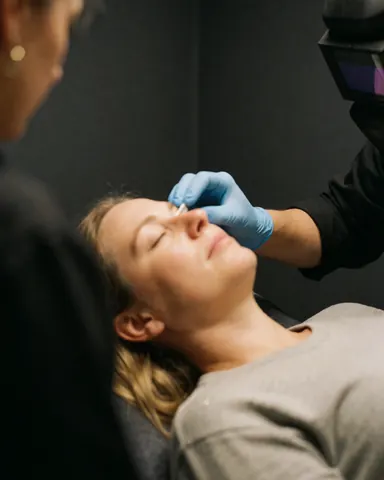 Cliente les yeux fermés recevant des soins post-microblading, technicienne en gant bleu appliquant un produit près de l'œil dans un salon de beauté.
