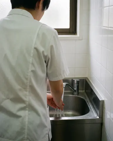 Un praticien de shiatsu se lave les mains dans un évier en acier inoxydable, avec des murs carrelés blancs et une lumière naturelle entrant par une fenêtre.
